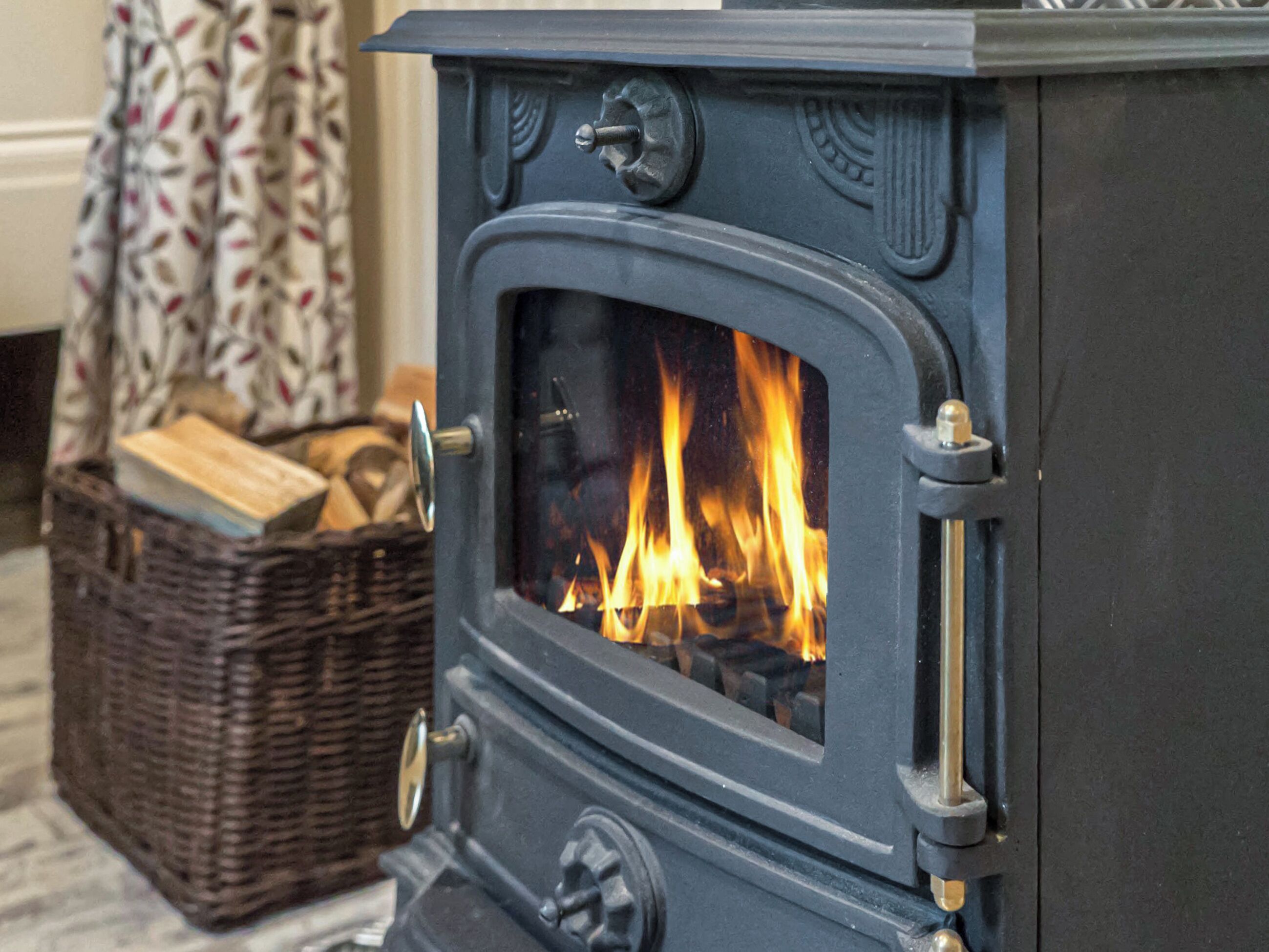 Un poêle à bois avec du feu à l’intérieur, un panier en osier rempli de bois de chauffage et, au fond, un rideau à motifs.