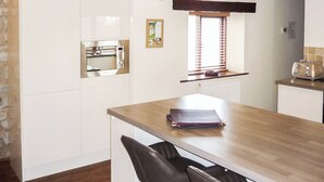 A compact kitchen with white cabinetry, a built-in microwave, a window with blinds, and a wooden table with a book. - This delightful single storey cottage offers cosy holiday accommodation. (Kirkbymoorside)