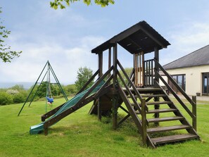 A playground with a slide and swing set, wooden climbing structure, and a house in the background. - 3 bedroom accommodation in Dunterton, near Tavistock (Dunterton, near Tavistock)