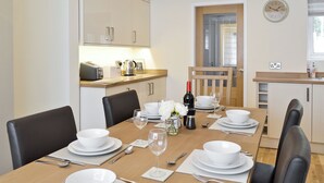 Een eethoek met een houten tafel voor vier personen, witte borden, kommen en bestek. Er hangt een klok aan de muur en er is een raam met jaloezieën.