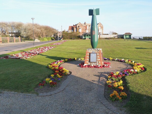 Un monument commémoratif composé d’un obélisque vert, entouré de parterres de fleurs et d’un chemin piétonnier dall é.