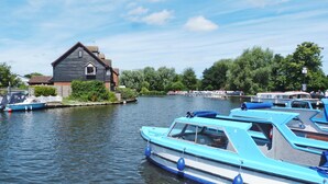 Een rustige rivier met aangemeerde boten, een huis met schuin dak en weelderige begroeiing op de achtergrond.