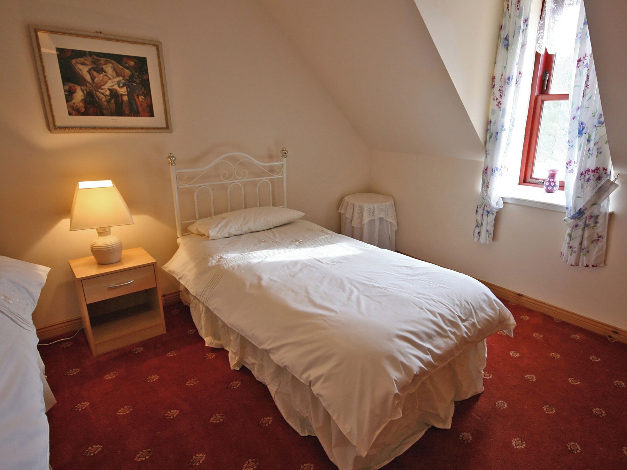 A bedroom with a single bed, a bedside table with a lamp, a small round table, a window with floral curtains, and a painting on the wall.