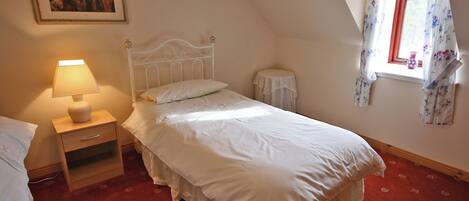 A bedroom with a single bed, a bedside table with a lamp, a small round table, a window with floral curtains, and a painting on the wall.