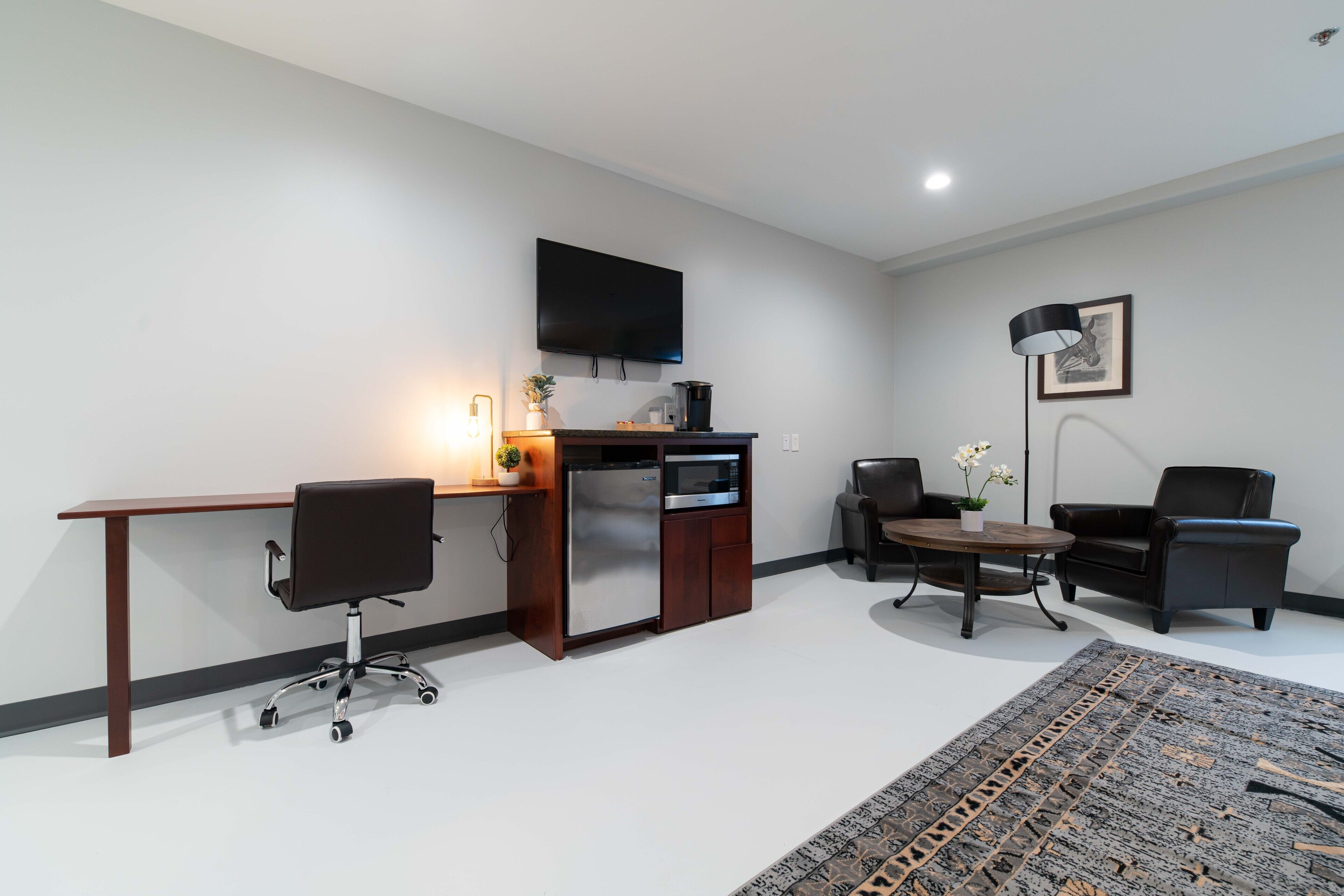 A modern living room with a wooden desk, a built-in microwave, a flat-screen TV, a coffee machine, two black leather chairs, a round coffee table, and a patterned rug.