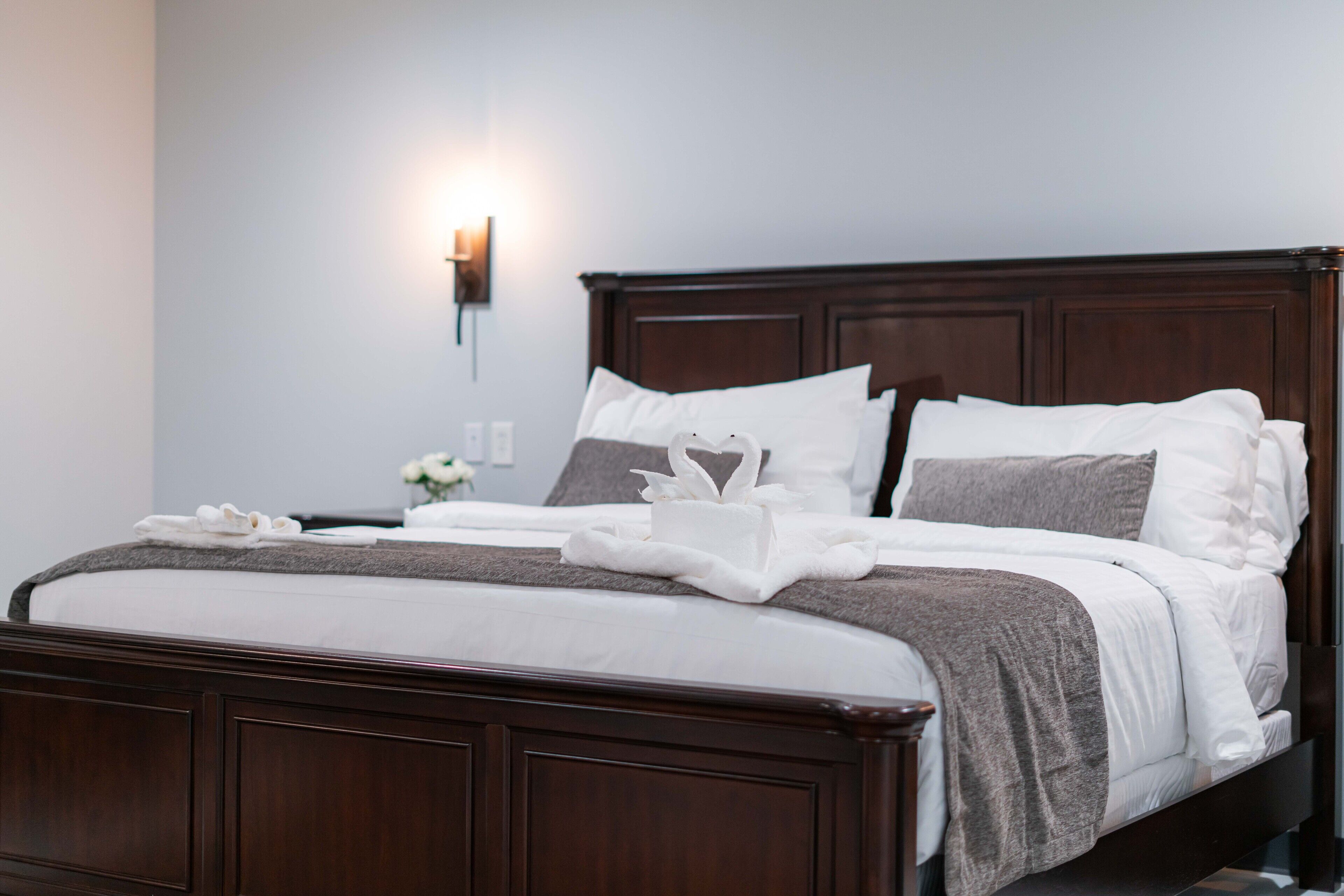A neatly made bed with white linens, a brown headboard, and a wall-mounted light fixture.
