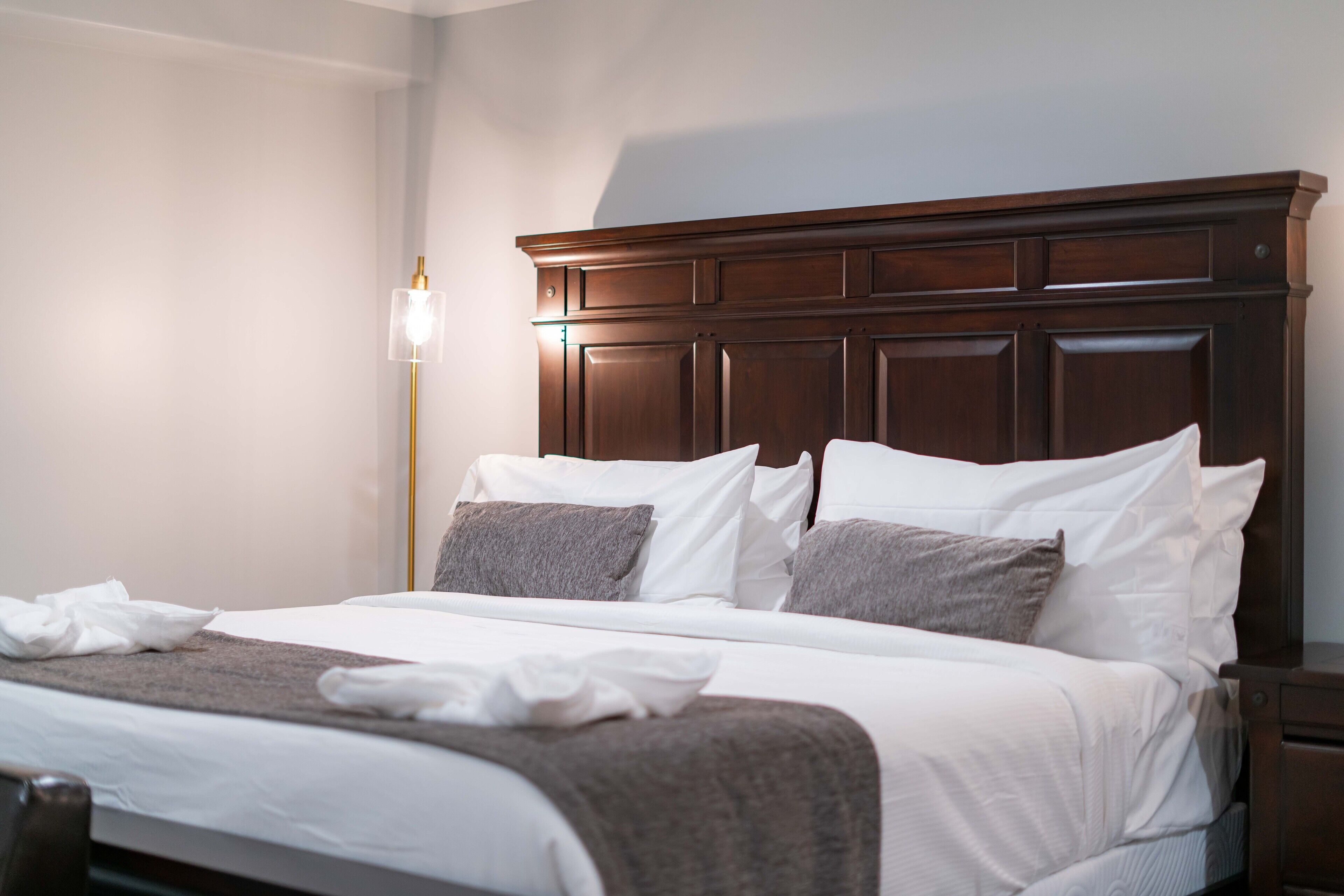 Cama bem arrumada com roupa de cama branca e cinza, cabeceira de madeira e abajur.