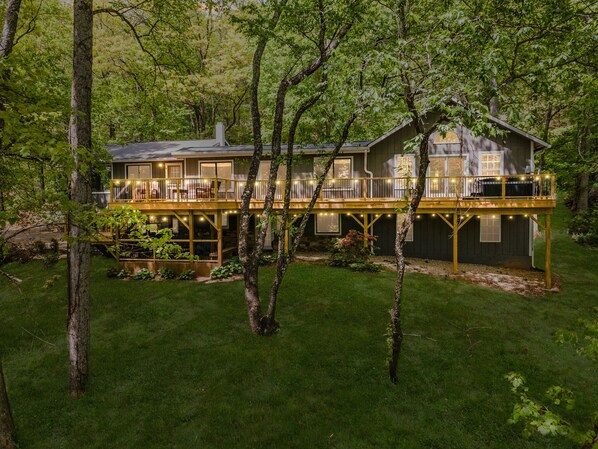 Une maison à deux étages dotée d’une terrasse et de nombreuses fenêtres, entourée d’arbres.
