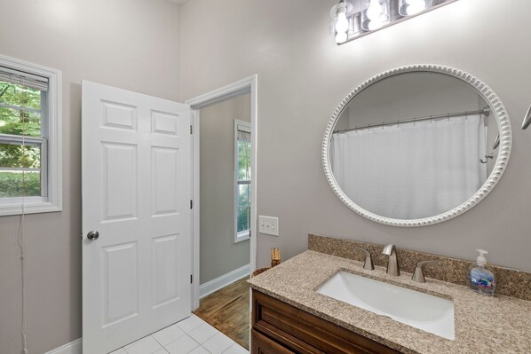A bathroom with a white door, a round mirror, a sink with a granite countertop, and a shower curtain.