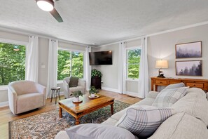 Un salon spacieux avec un grand canapé, une table basse et un téléviseur fixé au mur. De grandes fenêtres laissent entrer la lumière naturelle et offrent une vue sur la verdure environnante.