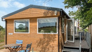 Una cabaña de madera con dos ventanas grandes, un patio con mesa y sillas, y una reja metálica.