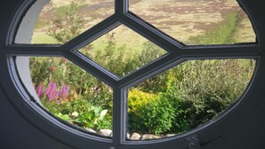 Une fenêtre ornée d’un motif en losanges, à travers laquelle on aperçoit un paysage verdoyant et fleuri.
