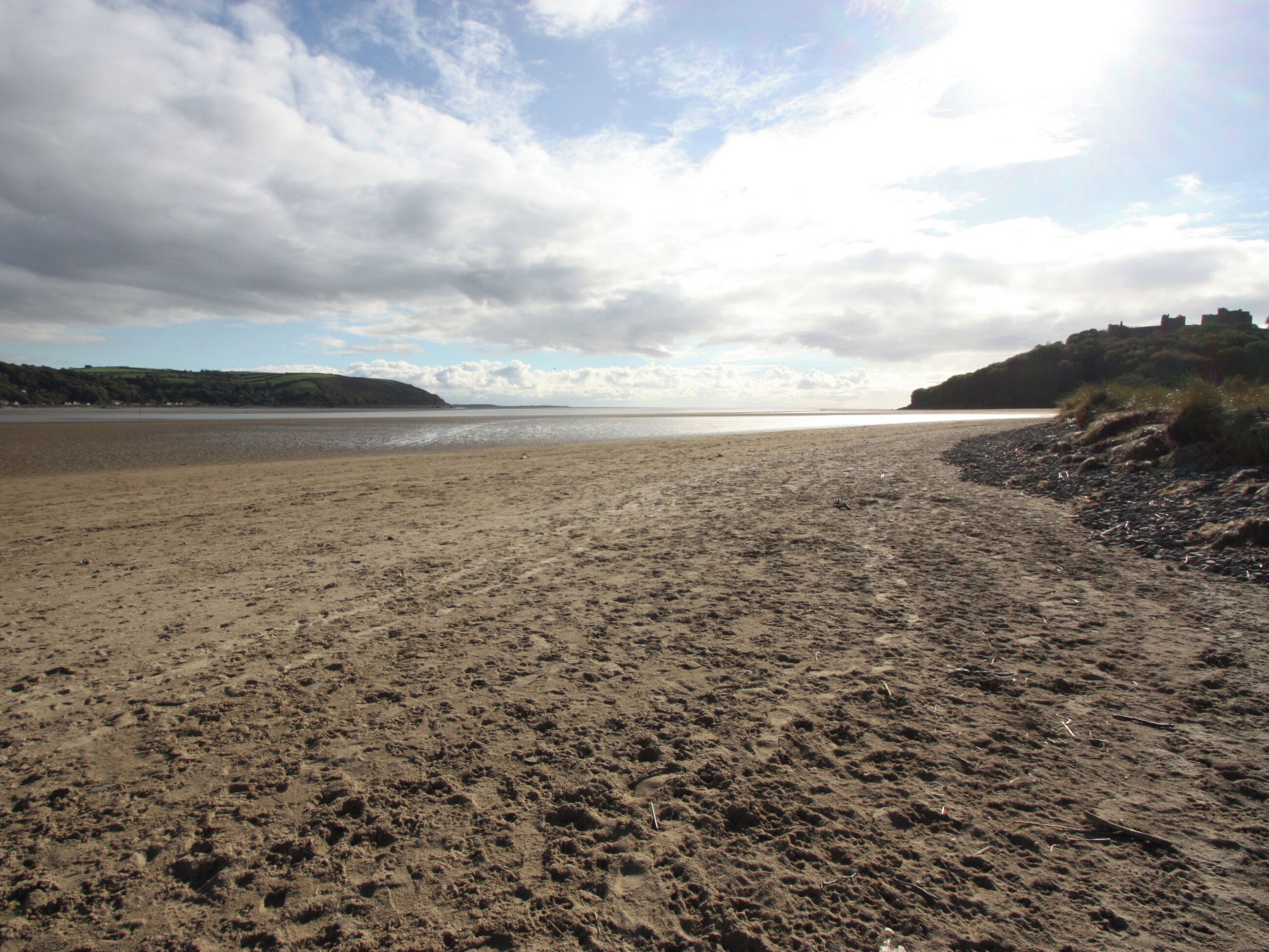 Une vaste plage de sable fin, avec des vagues douces, un ciel dégagé et des collines à l’horizon.