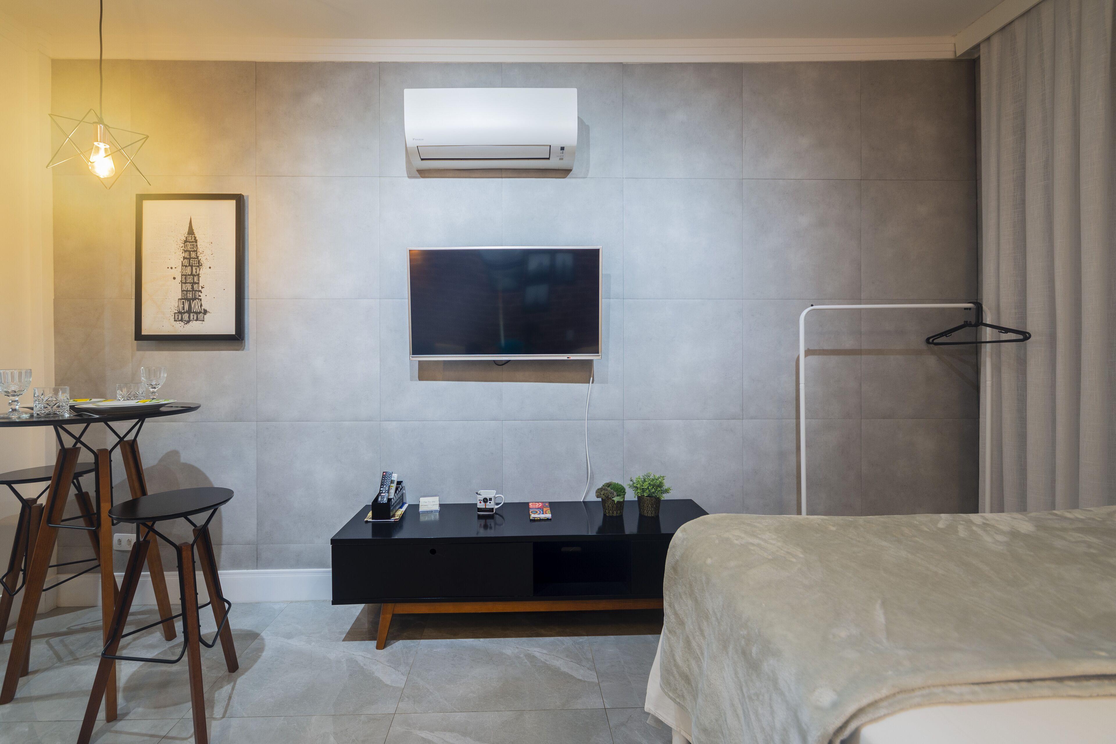 A modern hotel room with a bed, a TV mounted on the wall, a desk with chairs, and a framed picture.