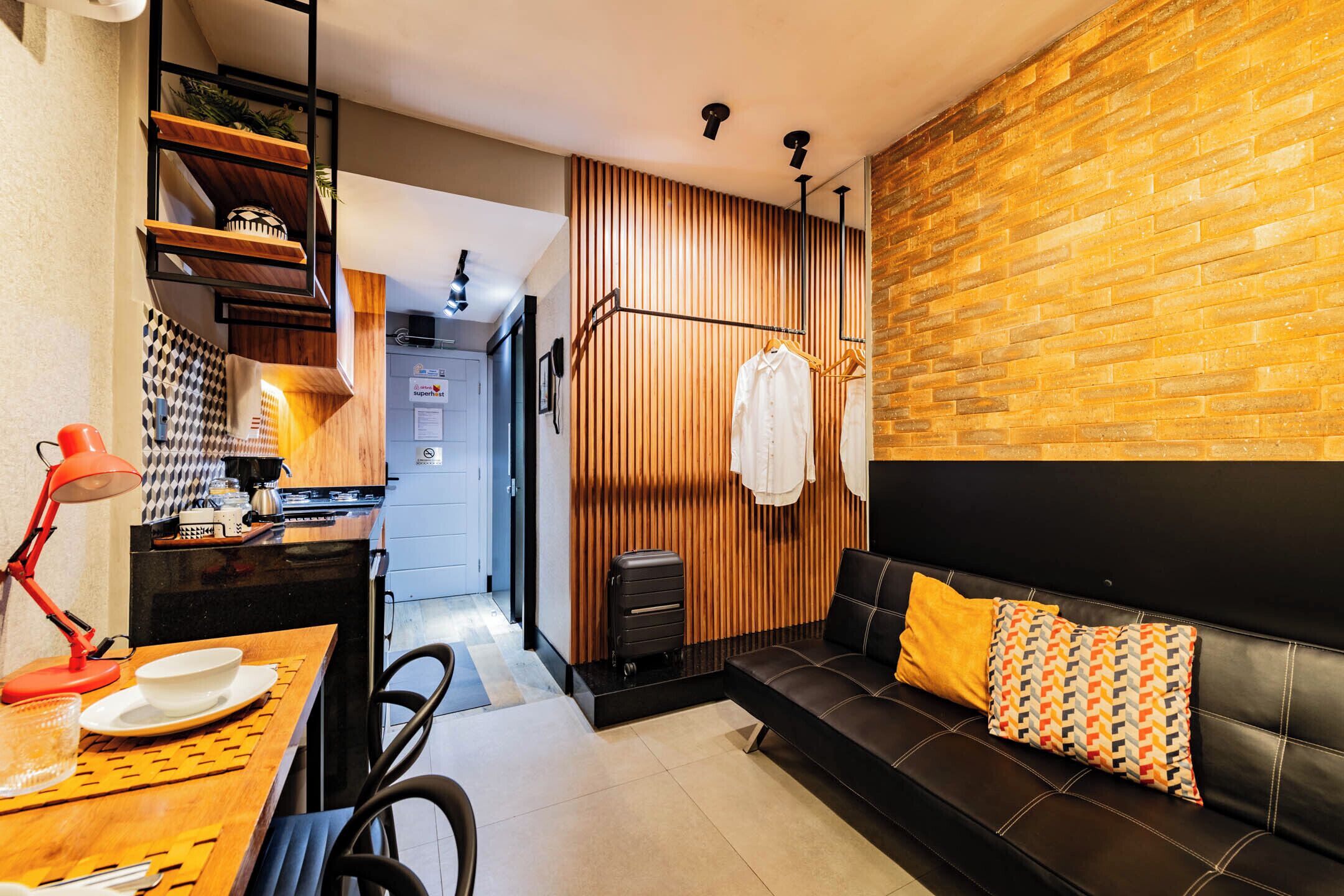 Quarto de hotel moderno com parede de destaque em madeira, sofá de couro preto, área de refeições com mesa e cadeiras, e um cabideiro suspenso.