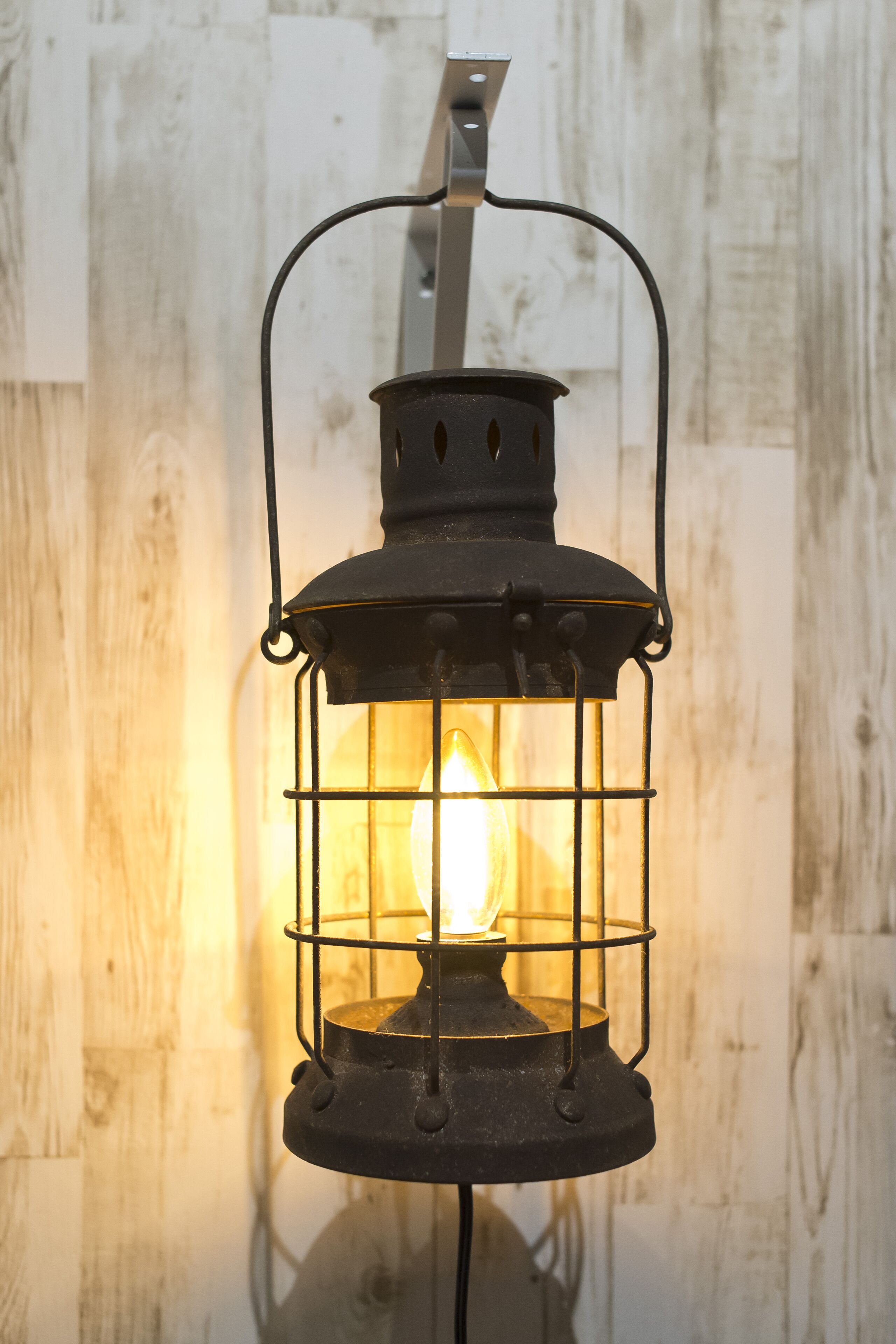 A vintage-style lantern with a visible light source inside, mounted on a wall with a rustic wooden texture.