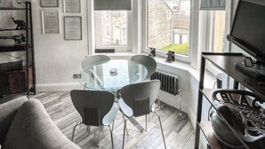Un salon moderne avec une table et des chaises en verre, un canapé, un téléviseur posé sur un meuble et une fenêtre donnant sur des immeubles.