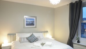 Una cama bien hecha con sábanas blancas y dos almohadas, un cuadro en la pared, una ventana con cortinas y una lámpara de noche.