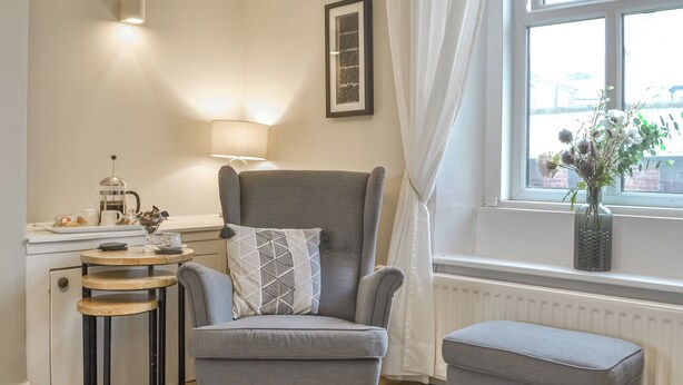Una sala moderna con un sillón gris, una mesita redonda pequeña, una mesa de centro y una ventana con cortinas.