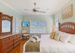 A bedroom with a bed, a wooden dresser, a chair, a ceiling fan, and a window with blinds.
