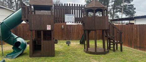 Una estructura de juego de madera con un tobogán y zonas de escalada, rodeada por una valla de madera.