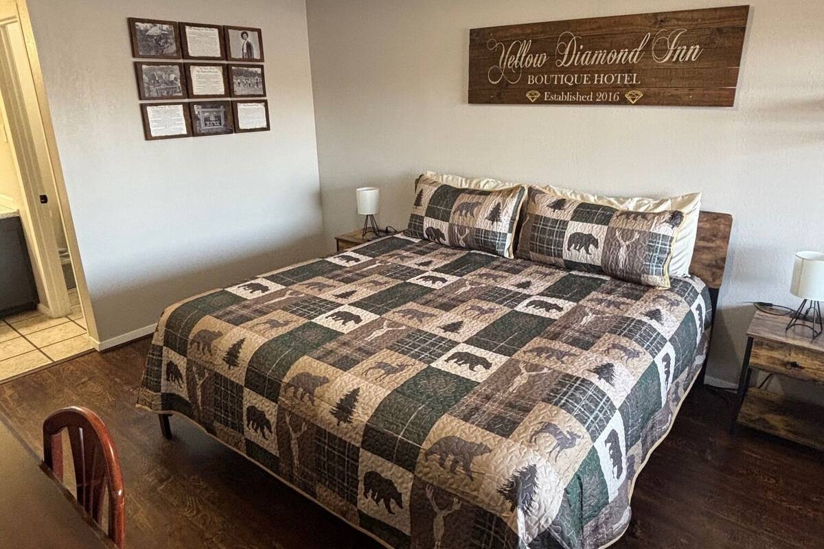 Quarto com cama grande, cabeceira de madeira, mesa de cabeceira com abajur, placa na parede e colagem de fotos emolduradas.