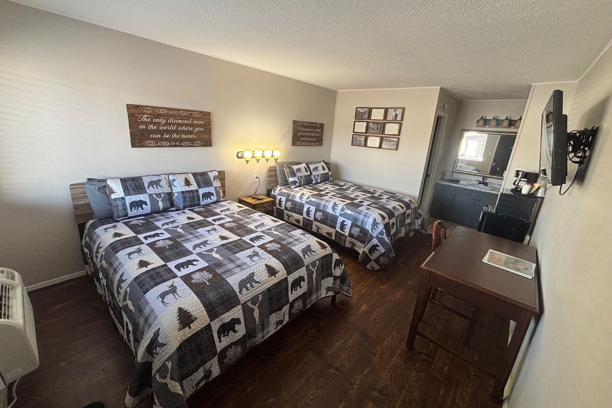 Quarto com duas camas, uma escrivaninha e um espelho. Possui decorações nas paredes e um ventilador de teto.