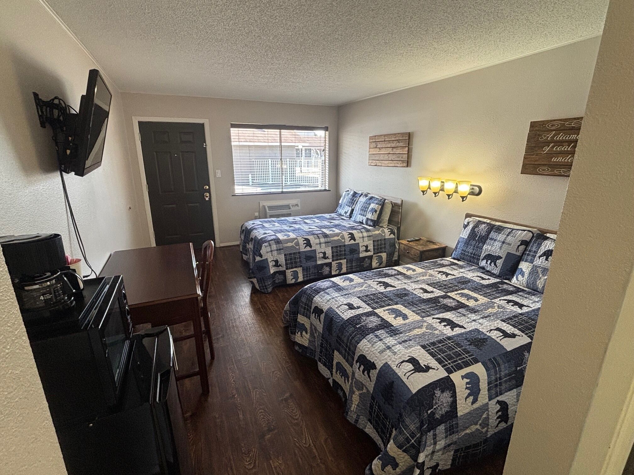 A small, cozy room with two beds, a desk, and a chair. There is a TV mounted on the wall, a window with blinds, and a wall-mounted light fixture.