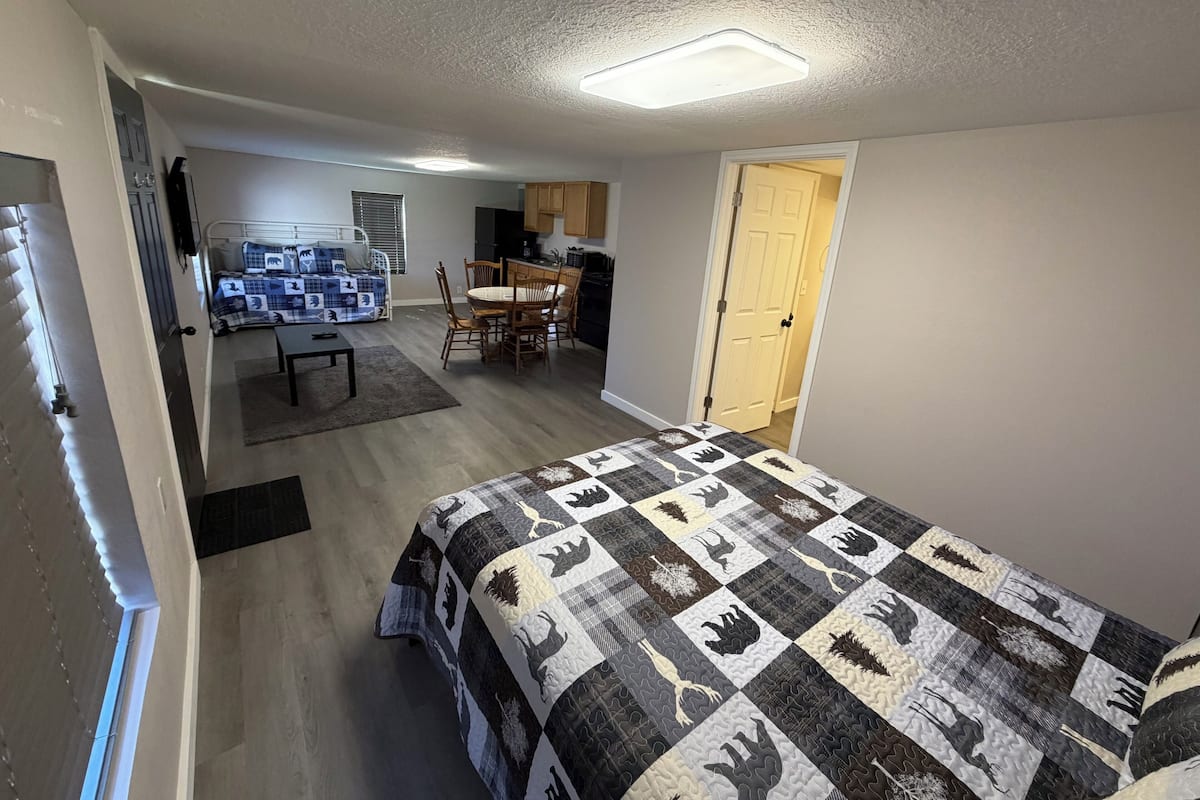 Quarto com cama, escrivaninha pequena, cadeira, mesa de jantar com cadeiras, cozinha e porta.