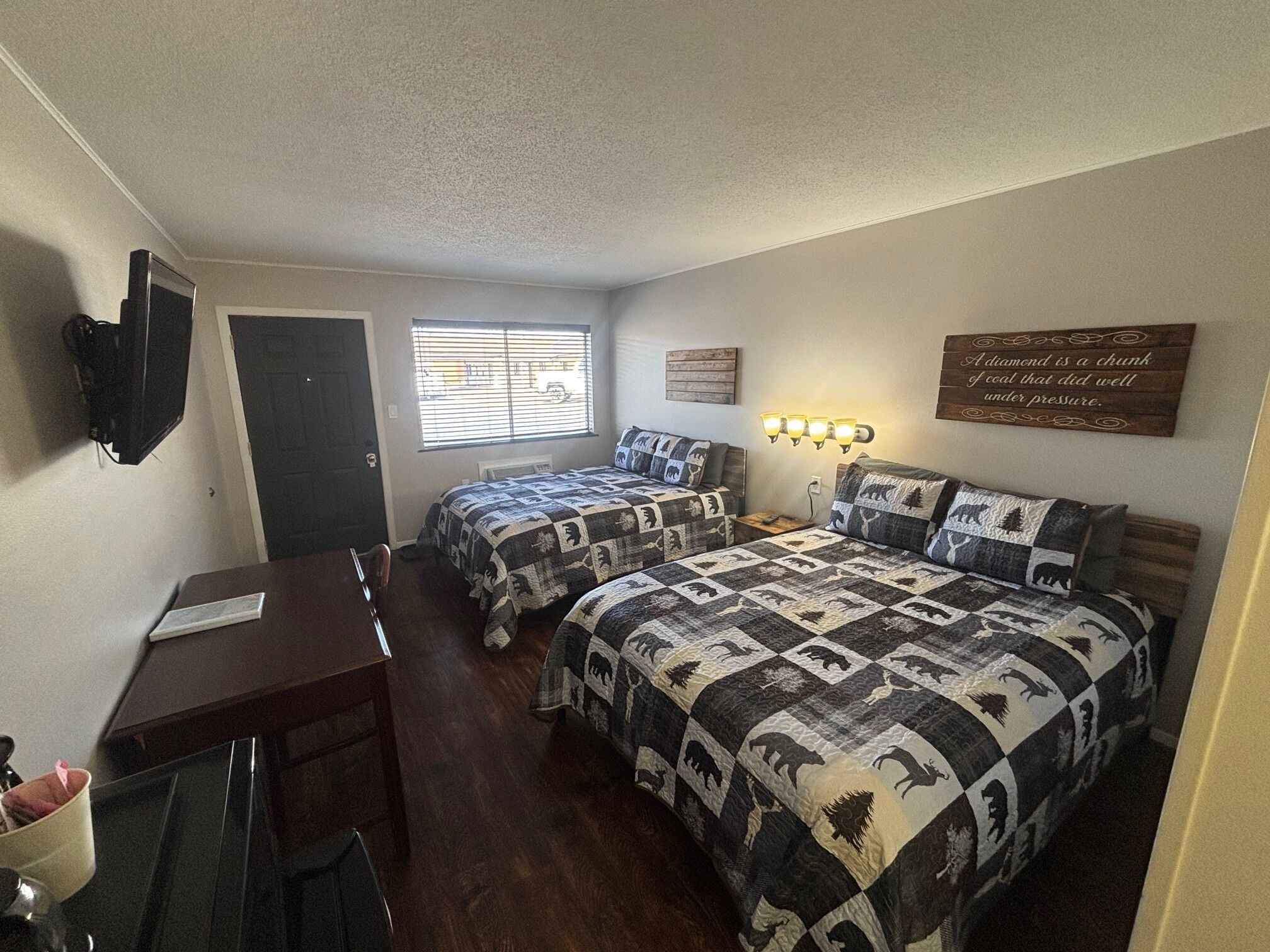 A bedroom with two beds, a desk, a TV, a window with blinds, and a motivational quote on the wall.
