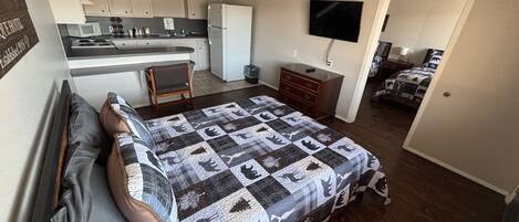 Una habitación pequeña con cama, escritorio, televisión y kitchenette.