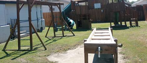 A backyard with a swing set, a slide, a playhouse, and a wooden table with a hose.