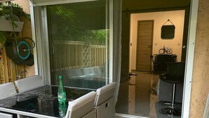 Un patio exterior con puerta de vidrio que da a una habitación, muebles de mimbre y una botella verde sobre la mesa.