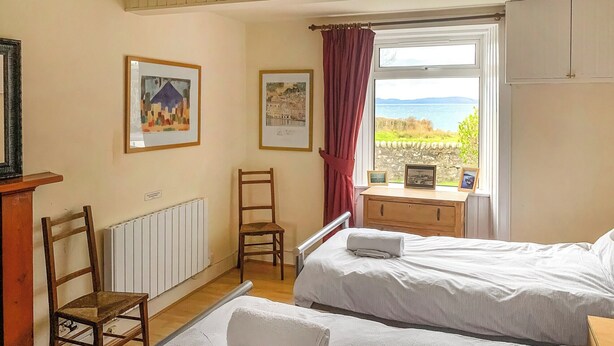 A bedroom with two beds, a wooden chair, a radiator, a window with a view of the sea, and framed pictures on the wall.