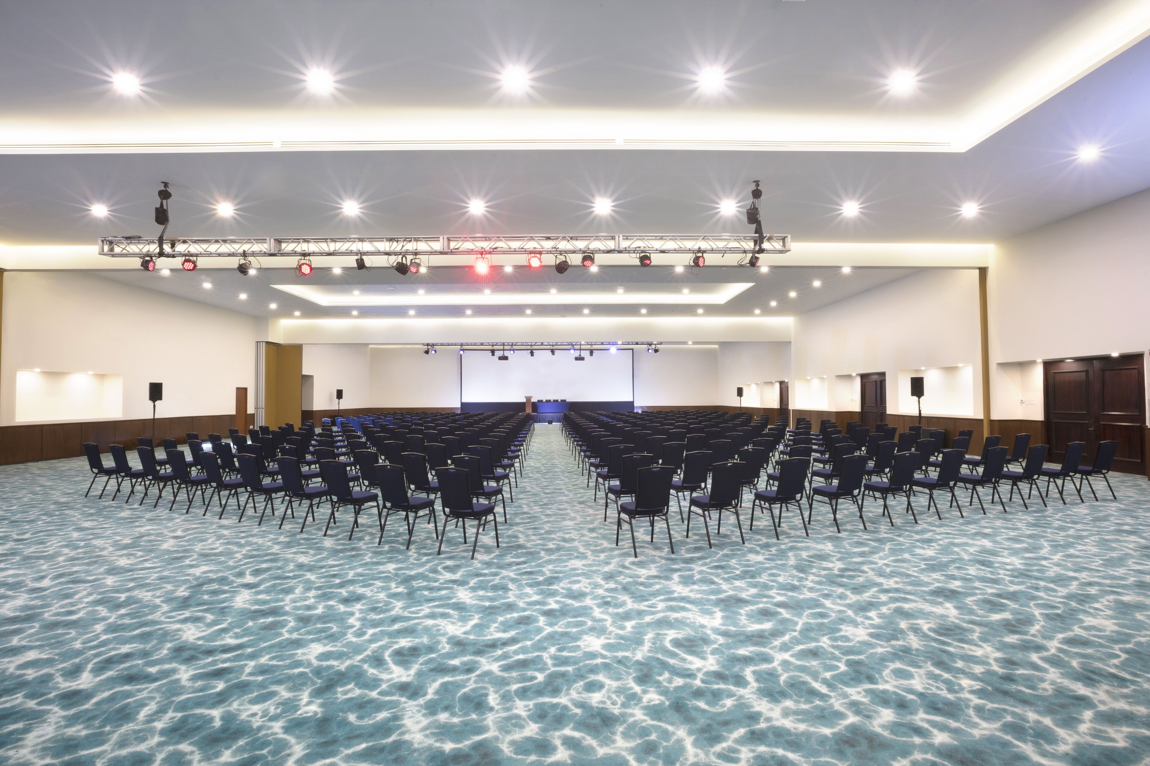 Un salón de conferencias amplio con filas de sillas, un escenario con iluminación y una pantalla de proyección.
