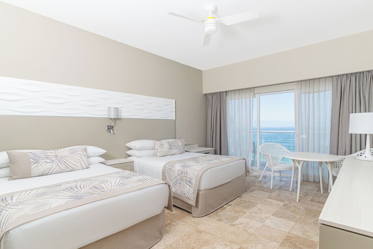 A hotel room with two beds, a desk, a chair, a ceiling fan, and a view of the ocean.