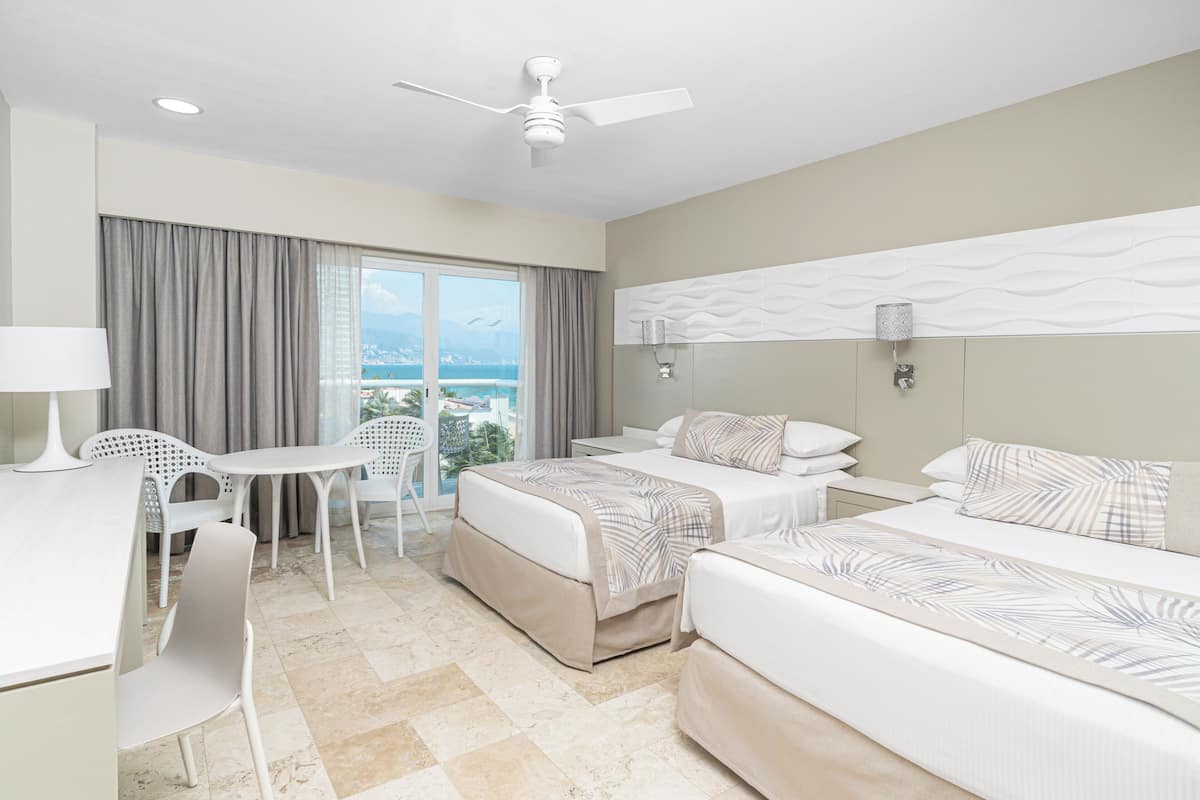 A hotel room with a bed, a desk, a chair, a ceiling fan, and a view of the ocean.