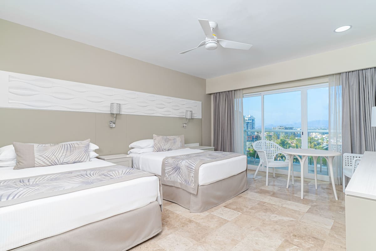 A hotel room with two beds, a ceiling fan, a balcony with a table and chairs, and a city view.