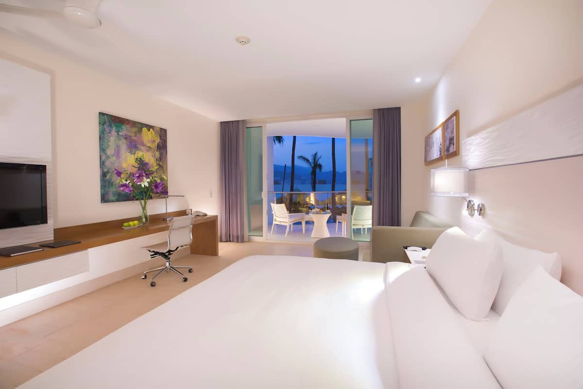 A modern hotel room with a large bed, a desk with a chair, a TV, a painting, and a balcony with a view of palm trees and the ocean.