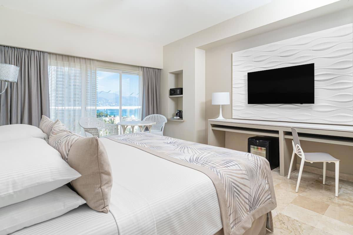 A hotel room with a bed, a desk, a chair, a TV, and a view of the city.