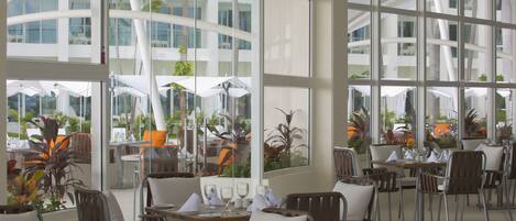 Ein modernes Restaurant mit großen Fenstern, Zimmerpflanzen und ordentlich angerichteten Tischen und Stühlen.