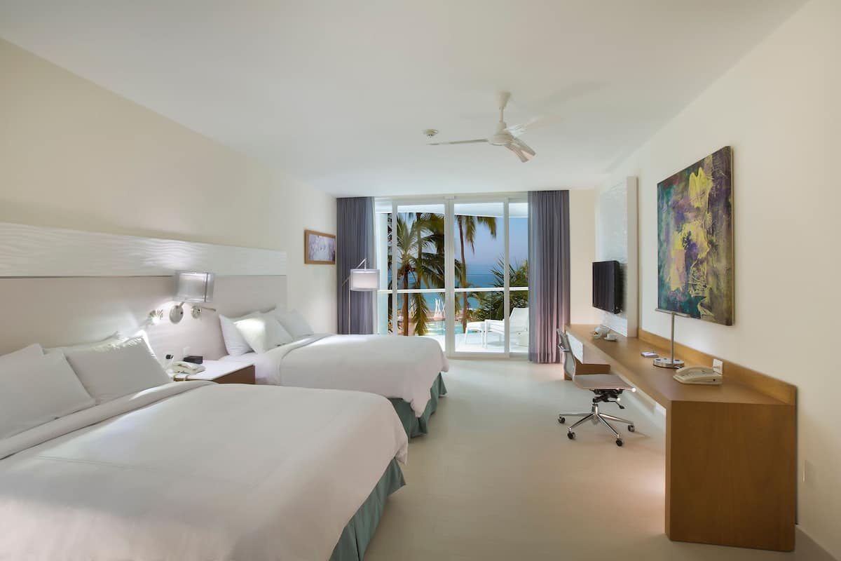 A hotel room with two beds, a desk, a chair, a ceiling fan, and a view of palm trees and the ocean.