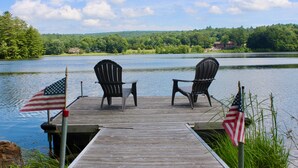 A wooden dock with two Adirondack chairs and American flags, overlooking a calm lake with a forested shoreline. - Discover Your Perfect Getaway! Beautiful Scenic Lakefront Condo (Montague)