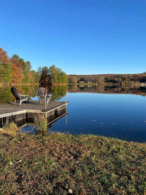 Un muelle con dos sillas Adirondack, un lago tranquilo y árboles de colores otoñales al fondo.