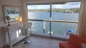 Una habitación con un ventanal grande con vistas a un lago, un tocador blanco con espejo, un sofá rojo y una pequeña mesa con lámpara.