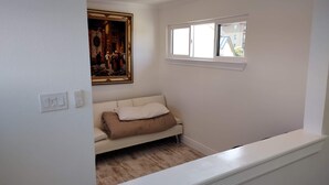 Una habitación con pared blanca, ventana, cuadro y sofá color beige con un cojín.