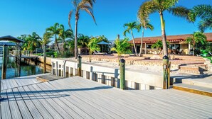 A wooden dock leading to a sandy beach area with palm trees and a building with a red roof.