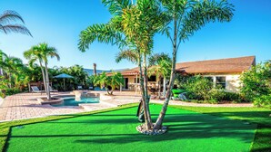 A pool area with artificial grass, palm trees, and a building with a tiled roof.