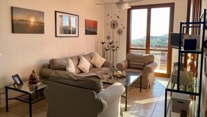 Un salon comprenant un canapé, une table basse et une vitrine. Les murs sont ornés de tableaux encadrés, et une fenêtre offre une vue sur l’extérieur.
