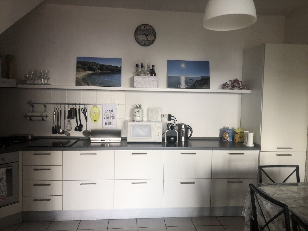Une cuisine avec des meubles blancs, un four à micro-ondes, une bouilloire et une horloge au mur.